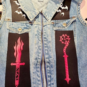 May include: A denim vest with black fabric patches featuring a flaming sword, a spiked chain, and a bat design. The vest has a zipper closure and a label that reads "Chaps by Chaps".