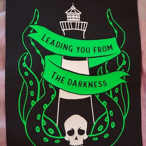 Può includere: Un cerotto di stoffa nero con un faro bianco e un polpo verde. Il faro ha un teschio alla base e uno striscione che recita "Leading you from the darkness."