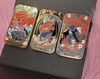 Puede incluir: Tres pines de esmalte de color dorado con imágenes de cartas de tarot. Los pines representan las cartas El Loco, Templanza y Muerte. Cada pin tiene un esquema de color negro y rojo con detalles dorados.