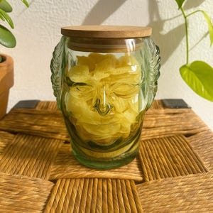 Puede incluir: Un tarro de cristal transparente con diseño de rostro, lleno de objetos amarillos y coronado con una tapa de madera. El tarro está sobre una superficie tejida, con plantas verdes al fondo. El tarro mide unos 15 cm de alto.