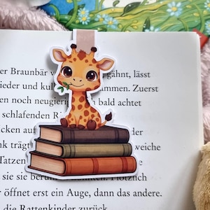 Könnte beinhalten: Ein bezaubernder Lesezeichen mit einer Cartoon-Giraffe mit braunen Flecken, die auf einem Stapel von drei Büchern sitzt. Die Giraffe hält ein grünes Blatt im Mund. Das Lesezeichen befindet sich in einem Buch mit deutschem Text.