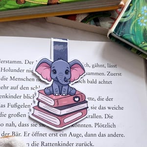 Könnte beinhalten: Ein bezaubernder Lesezeichen mit einem Cartoon-Elefanten mit rosa Ohren, der auf einem Stapel Bücher sitzt. Das Lesezeichen befindet sich in einem offenen Buch mit deutschem Text. Das Design ist blau, rosa und weiß.