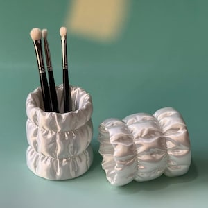 May include: White satin makeup brush holder with three brushes and a matching accessory. The holder is cylindrical with three stacked sections. The accessory is a rounded shape with a similar design. The background is a solid teal colour.