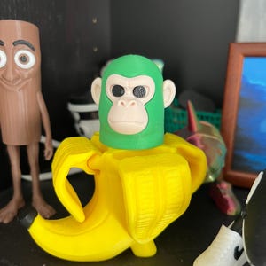 May include: A quirky 3D-printed sculpture featuring a monkey head with a green cap emerging from a yellow banana. The monkey has a tan face and black eyes. Other figurines are visible in the background.