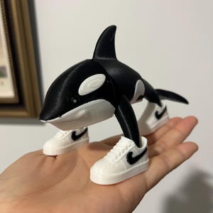 May include: A 3D-printed orca figurine in black and white, wearing miniature white sneakers with black swoosh logos. The orca has a dorsal fin and white markings around its eyes.