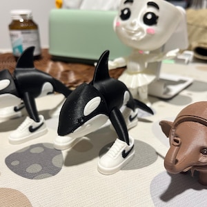 Op de afbeelding: Een verzameling 3D-geprinte beeldjes, waaronder twee zwart-witte orka's die miniatuur witte sneakers met zwarte swooshes dragen. Er is ook een bruin, olifantachtig figuur en een wit, lachend gezichtsvormig object.