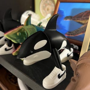 Op de afbeelding: Een zwart-wit orka beeldje met een witte sneaker aan de voet. De orka staat op een plank met andere beeldjes, waaronder een haai en een schildpad schilderij op de achtergrond. De sneaker heeft een zwarte Nike swoosh.