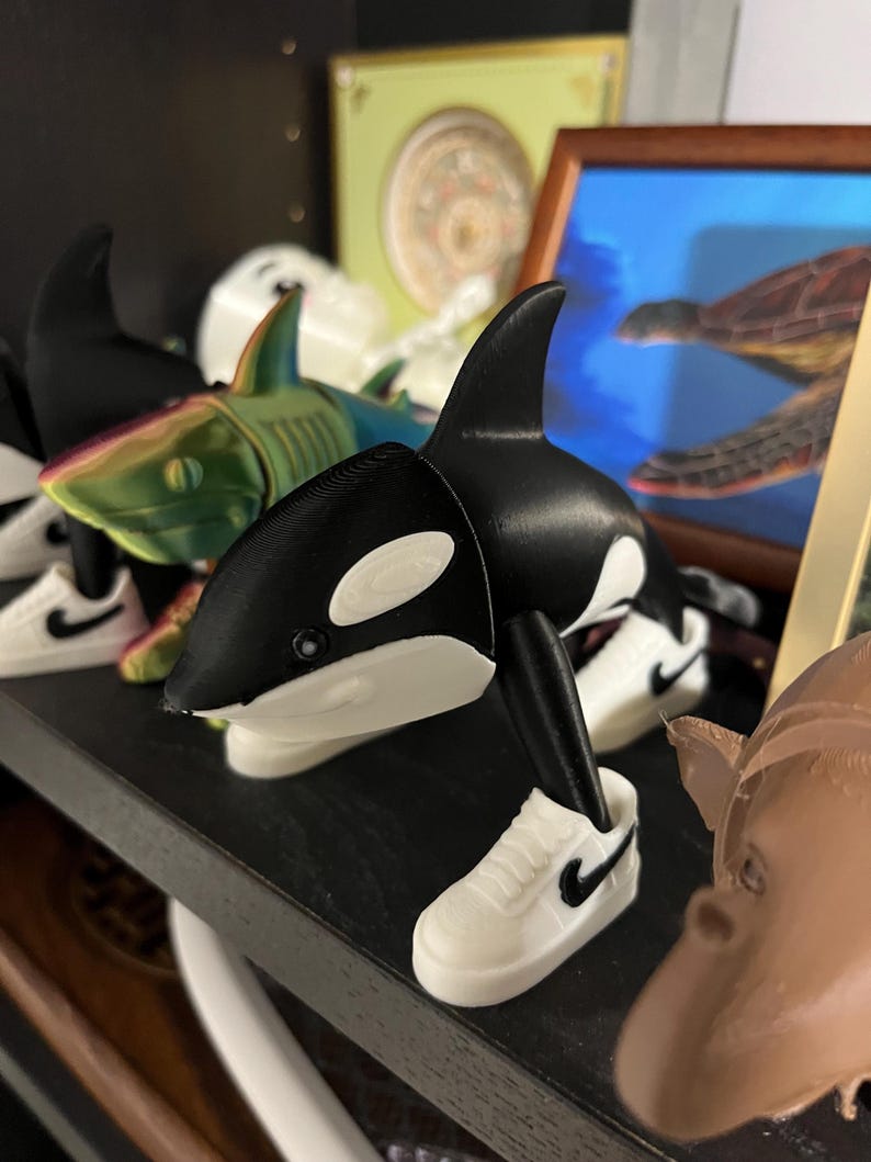 Op de afbeelding: Een zwart-wit orka beeldje met een witte sneaker aan de voet. De orka staat op een plank met andere beeldjes, waaronder een haai en een schildpad schilderij op de achtergrond. De sneaker heeft een zwarte Nike swoosh.