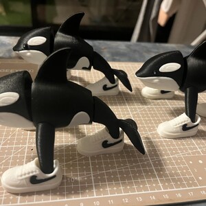 Op de afbeelding: Drie zwart-witte orca-beeldjes die elk kleine witte sneakers met zwarte swoosh-logo's dragen. De orka's hebben een speels ontwerp, met hun vinnen en staarten zo gepositioneerd alsof ze in beweging zijn. De beeldjes worden op een patroonoppervlak weergegeven.