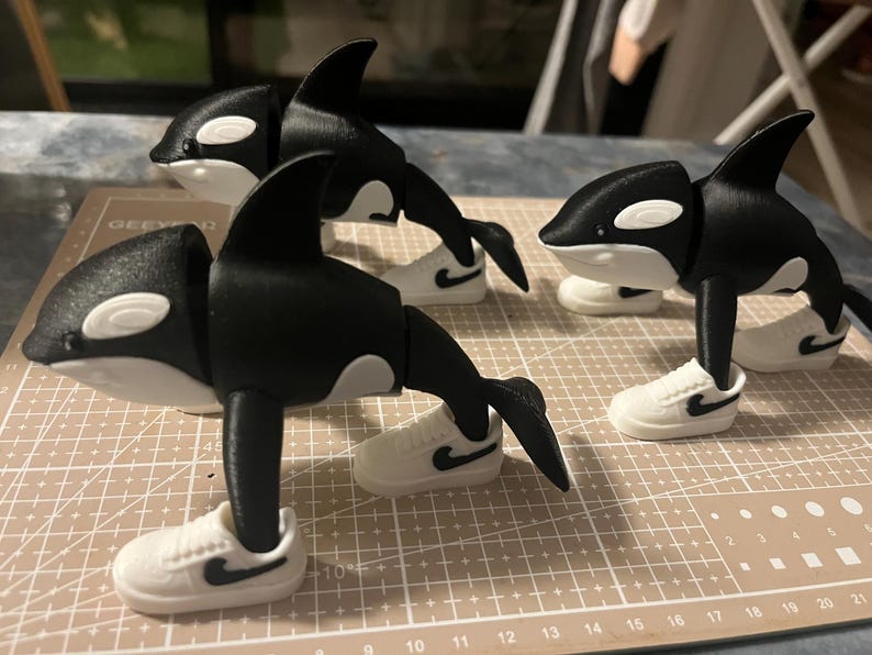 Op de afbeelding: Drie zwart-witte orca-beeldjes die elk kleine witte sneakers met zwarte swoosh-logo's dragen. De orka's hebben een speels ontwerp, met hun vinnen en staarten zo gepositioneerd alsof ze in beweging zijn. De beeldjes worden op een patroonoppervlak weergegeven.