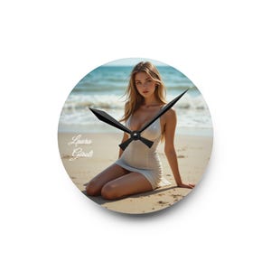 May include: Round wall clock featuring a beach scene with a woman in a white dress. The clock hands are black. The clock face includes the text "Laura Giroti".