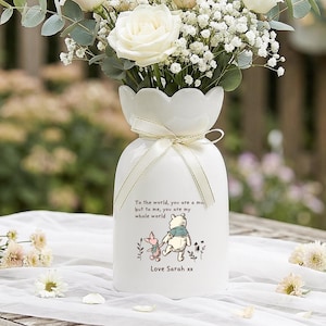Pode incluir: Um vaso de cerâmica branca com a parte superior ondulada, amarrado com uma fita, segurando um buquê de rosas brancas e rosa, gipsofila e vegetação. O vaso apresenta uma ilustração de Winnie the Pooh e o texto "To the world, you are a mom, but to me, you are my whole world. Love Sarah xx."