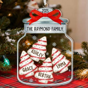 May include: A clear, jar-shaped Christmas ornament with a red bow and the year 2025. Inside are four tree-shaped cookies with names: Grace, Ashley, Bertha, and Emma. The text "THE RAYMOND FAMILY" is above the cookies.