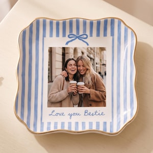 May include: A square ceramic trinket dish with wavy edges and a gold-painted rim. The dish features a light blue and white vertical striped pattern, a blue bow graphic, and a central photograph of two people holding coffee cups with the text Love you Bestie.