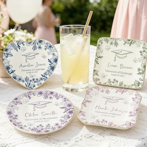May include: Graduation-themed ceramic plates in various shapes, adorned with floral designs and personalized text. Each plate features a graduation cap illustration and the graduate's name, university, and class year. A refreshing drink with ice and a straw is also visible.