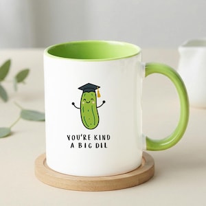 May include: White ceramic mug with a green interior and handle. The mug features a cartoon pickle wearing a graduation cap and the text "YOU'RE KIND A BIG DIL". The mug sits on a wooden coaster.
