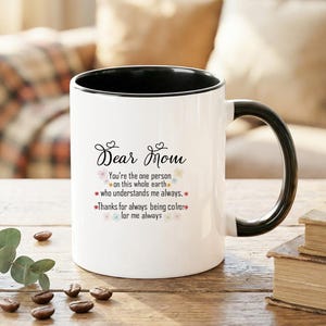 Könnte beinhalten: Weißer Keramikbecher mit schwarzem Innenraum und Henkel. Der Becher trägt den Text "Dear Mom" in Schreibschrift, zusammen mit einer herzlichen Botschaft und floralen Akzenten. Der Becher steht auf einer Holzoberfläche.
