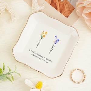 May include: A square white ceramic trinket dish with a gold-trimmed scalloped edge. The center features two floral illustrations with the names Tanner and Dallas written vertically, and the text IF FRIENDS WERE FLOWERS I WOULD PICK YOU.
