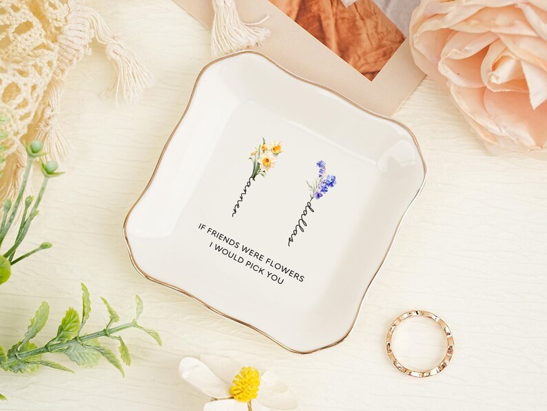 May include: A square white ceramic trinket dish with a gold-trimmed scalloped edge. The center features two floral illustrations with the names Tanner and Dallas written vertically, and the text IF FRIENDS WERE FLOWERS I WOULD PICK YOU.