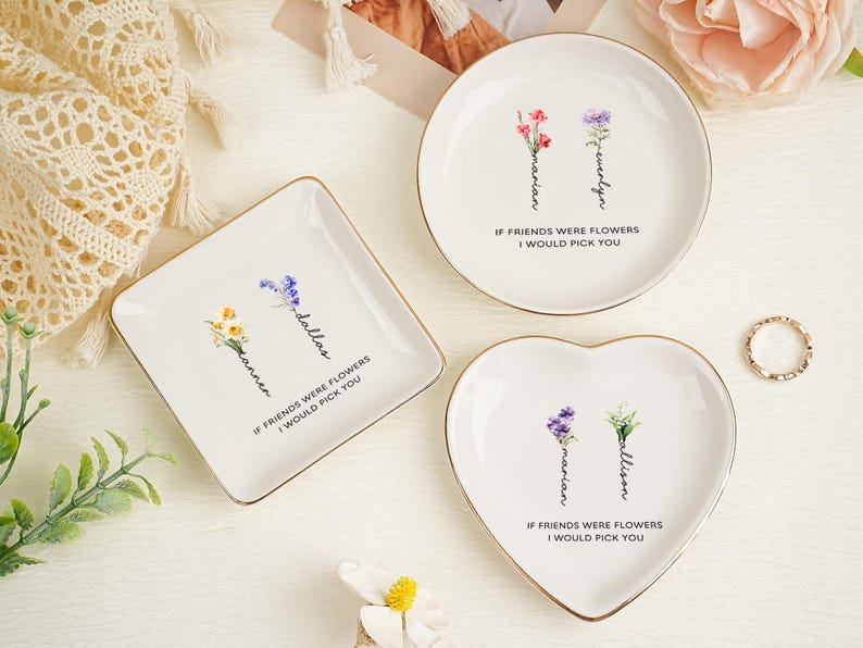 May include: Three white ceramic trinket dishes with gold-toned rims in square, round, and heart shapes. Each dish features two colorful floral illustrations with names written vertically on the stems and the text IF FRIENDS WERE FLOWERS I WOULD PICK YOU.
