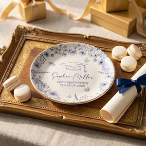 May include: A decorative plate with blue floral designs, a graduation cap, and the text "Sophie Miller, Cambridge University, CLASS OF 2026." The plate is on a gold-trimmed tray with macarons and a rolled diploma.