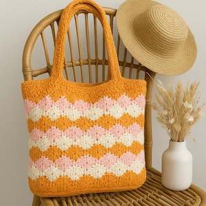 Könnte beinhalten: Eine gehäkelte Tragetasche in Orange, Rosa und Creme mit Zickzackmuster. Die Tasche hat zwei Henkel und steht auf einem Holzstuhl. Ein Strohhut und eine Vase mit Trockenblumen sind ebenfalls zu sehen. Ein modisches Accessoire.