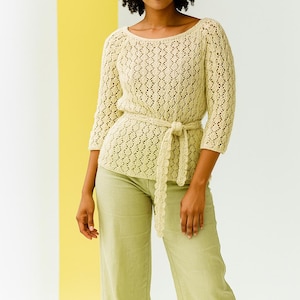 Könnte beinhalten: Ein hellbeige, offen gewebtes Strickoberteil mit Dreiviertelärmeln und einem passenden Gürtel, der in der Taille gebunden ist. Das Oberteil wird mit hellgrünen Hosen kombiniert. Der Hintergrund zeigt einen weißen und gelben Farbblock.