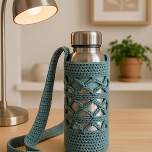 Peut inclure: Une gourde en acier inoxydable avec une housse et une sangle en crochet turquoise. La housse présente un motif en treillis. La bouteille est sur une surface en bois, avec une lampe et une image encadrée en arrière-plan.