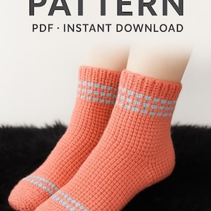 May include: Pair of coral-coloured crochet socks with grey detailing. The socks feature a textured design and a band of grey squares at the top. The image also includes the text "CROCHET SOCKS PATTERN PDF • INSTANT DOWNLOAD".