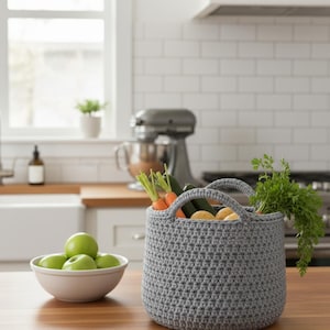 May include: A gray crocheted basket filled with fresh vegetables, including carrots, zucchini, and potatoes. The basket has two handles and sits on a wooden countertop. A bowl of green apples is in the foreground.