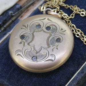 May include: A gold-colored, round pendant necklace with an ornate engraved design and small blue gemstones. The pendant is attached to a delicate gold chain. The necklace is displayed on a dark blue velvet surface, suggesting a vintage or antique piece.