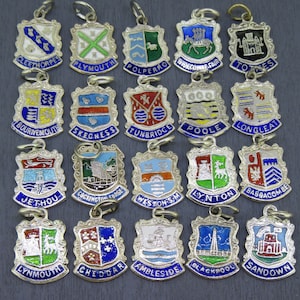 May include: A collection of 18 enamel and silver charms featuring coats of arms with the names of various towns and cities in the United Kingdom. The charms are in various colours, including blue, green, red, yellow, and white.