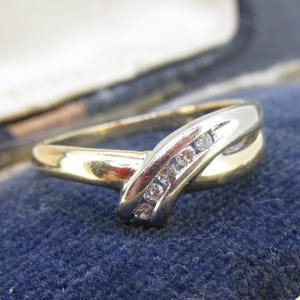 May include: A gold and silver-tone ring with a curved design. The ring features a row of small, clear gemstones set in the silver portion. The ring is displayed on a blue velvet surface.