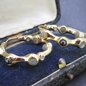May include: A pair of gold-toned hoop earrings with a unique design. The earrings feature a series of rounded, organic shapes, each adorned with small, iridescent gemstones. The earrings are displayed in a vintage jewelry box with a blue velvet interior.