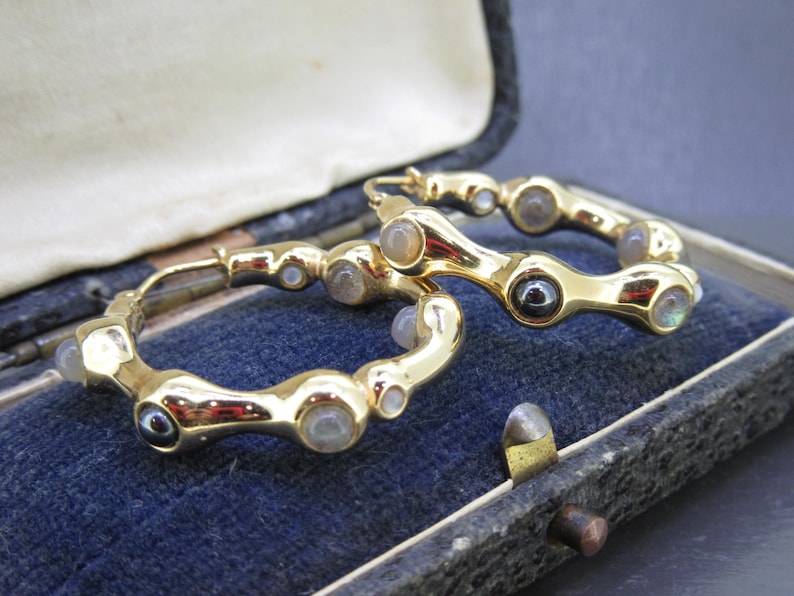 May include: A pair of gold-toned hoop earrings with a unique design. The earrings feature a series of rounded, organic shapes, each adorned with small, iridescent gemstones. The earrings are displayed in a vintage jewelry box with a blue velvet interior.