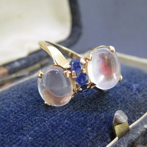 May include: A gold ring featuring three oval-shaped, translucent stones with a hint of color, accented by small blue gemstones. The ring is set against a blue velvet background, suggesting a jewelry display.