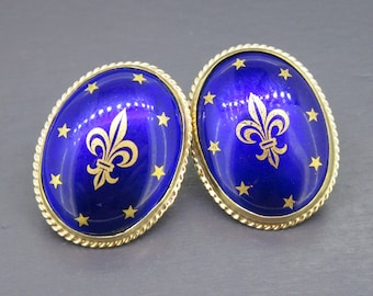 May include: A pair of oval-shaped earrings with a royal blue enamel surface. Each earring features a gold fleur-de-lis and star design, surrounded by a gold rope-like border. The earrings are set in a gold-toned metal.