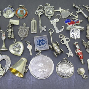 May include: A collection of silver charm pendants featuring various designs, including a graduation cap, a heart with a violin, a bell, a key, a Notre Dame logo, a church, a thermometer, a telephone, a bus, a golf ball, a pair of hands in prayer, and a variety of other designs.