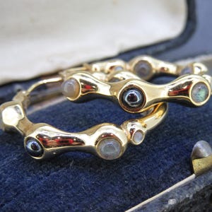 May include: Two gold-toned hoop earrings with a unique, linked design. Each earring features multiple small, round, iridescent gemstones. The earrings are displayed inside a vintage blue velvet jewelry box, showcasing their intricate details and craftsmanship.