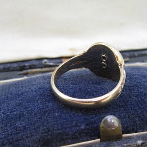 May include: A gold ring featuring a circular head with a dark, recessed center containing two small, vertically aligned circular metal studs. The interior of the band is engraved with the text OSTBY & BARTON.