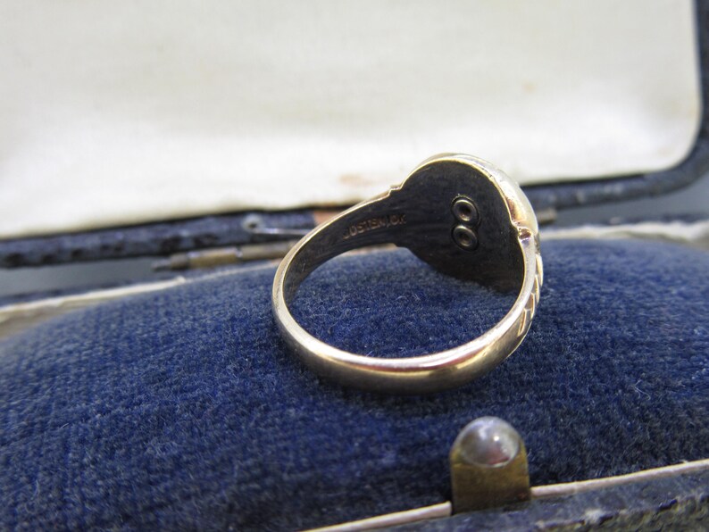 May include: A gold ring featuring a circular head with a dark, recessed center containing two small, vertically aligned circular metal studs. The interior of the band is engraved with the text OSTBY & BARTON.