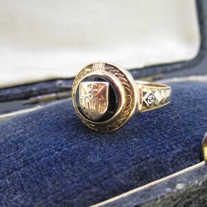 May include: A gold ring featuring a circular black onyx inlay centered with a metallic shield emblem. The band displays a diamond-shaped detail engraved with the number 69. The ring has a textured, decorative border surrounding the central stone.