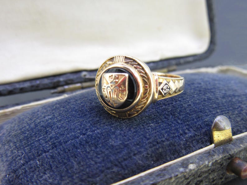 May include: A gold ring featuring a circular black onyx inlay centered with a metallic shield emblem. The band displays a diamond-shaped detail engraved with the number 69. The ring has a textured, decorative border surrounding the central stone.