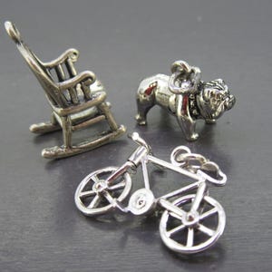 Pu&ograve; includere: Tre ciondoli in miniatura color argento: una sedia a dondolo, un bulldog e una bicicletta. La sedia a dondolo ha lo schienale a doghe. Il bulldog &egrave; in piedi. La bicicletta ha raggi dettagliati. Tutto su una superficie grigio scuro.