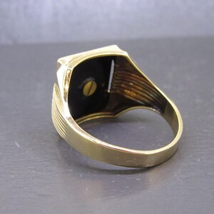 May include: A gold-toned ring with a rectangular black stone set in a gold frame. The ring has a classic design with a smooth band and detailed shoulders. The black stone is secured with a small screw.