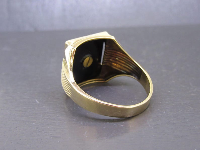 May include: A gold-toned ring with a rectangular black stone set in a gold frame. The ring has a classic design with a smooth band and detailed shoulders. The black stone is secured with a small screw.
