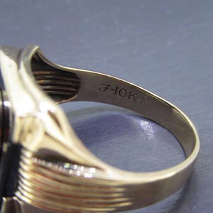 May include: A gold ring with a black rectangular stone and a circular emblem. The inside band is stamped with "7-10K-". The ring has a polished finish and a classic design, suitable for various occasions.
