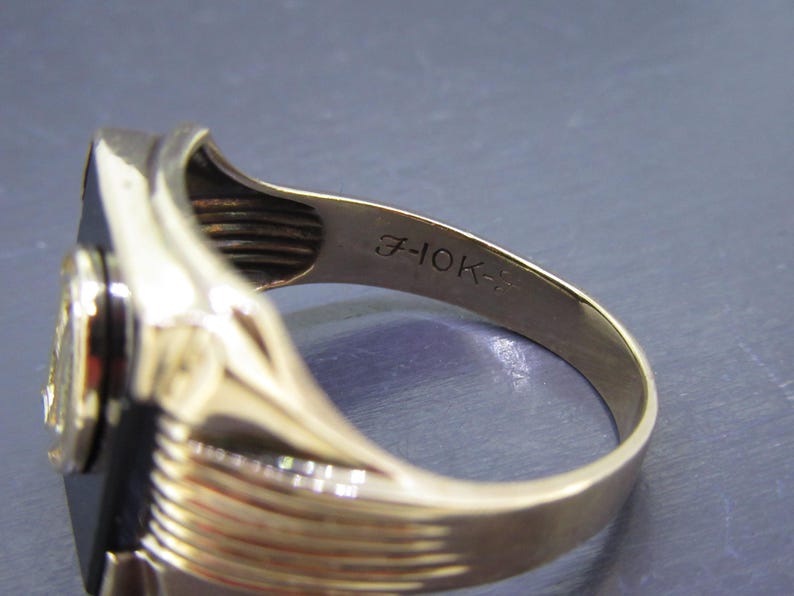 May include: A gold ring with a black rectangular stone and a circular emblem. The inside band is stamped with "7-10K-". The ring has a polished finish and a classic design, suitable for various occasions.