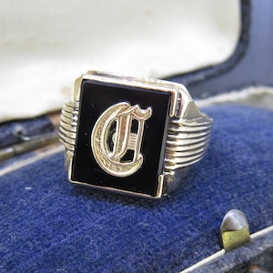 May include: A gold-toned ring with a black rectangular face. The face features a gold letter "C" in an ornate font. The ring has a ribbed design on the sides and is set against a blue velvet background.
