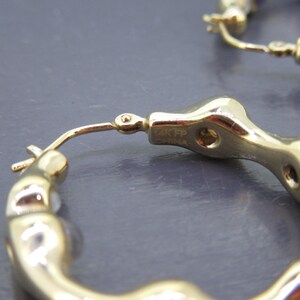 May include: Close-up of gold-colored hoop earrings. The earrings have a unique, textured design with a series of rounded, interconnected shapes. The earrings are marked with "14K FP CHINA". The background is a solid, dark gray color.